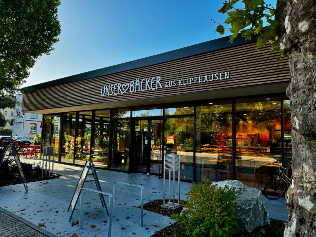 Ein schönes Gebäude mit Holzapplikationen und großen Fensterfronten, mit der Aufschrift "Unser Bäcker aus Klipphausen", darüber blauer Himmel. 