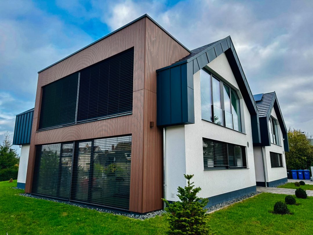 Ein modernes Doppelhaus bei bewölktem Himmel. Die linke Hälfte ist mit vertikalen braunen Holz- oder Holzimitat-Lamellen verkleidet und hat breite Fenster mit dunklen Außenjalousien. Die rechte Hälfte hat eine weiße Putzfassade mit einem dunklen Giebeldach und großen, dreieckigen Fenstern. Ein kleiner Nadelbaum steht im grünen Vorgarten.