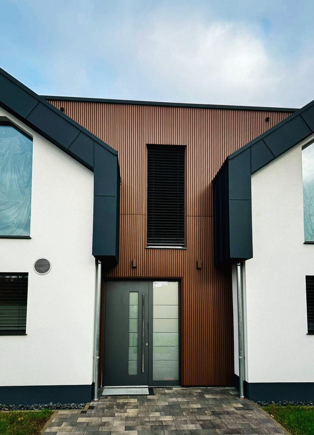 Der Eingangsbereich des modernen Doppelhauses. Er ist mittig zwischen den beiden weißen Giebelseiten platziert und komplett mit vertikalen braunen Holz- oder Holzimitat-Lamellen verkleidet. Eine moderne, dunkelgraue Eingangstür mit Glasausschnitten und ein schlichtes, hohes Fenster mit Jalousien sind zu sehen.