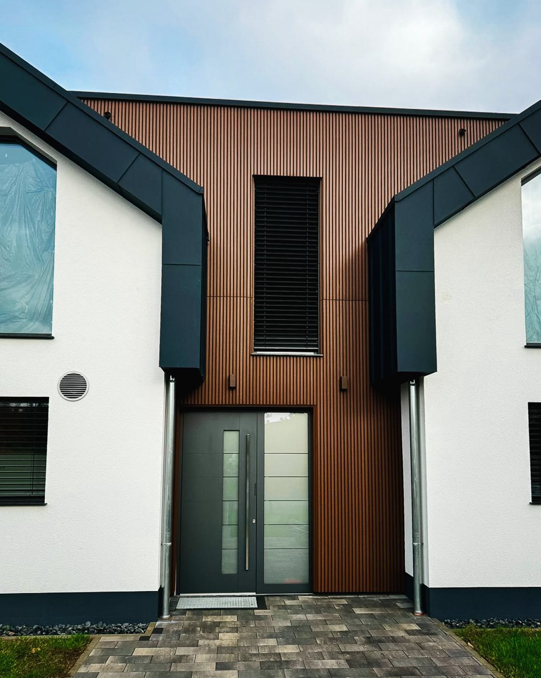 Der Eingangsbereich des modernen Doppelhauses. Er ist mittig zwischen den beiden weißen Giebelseiten platziert und komplett mit vertikalen braunen Holz- oder Holzimitat-Lamellen verkleidet. Eine moderne, dunkelgraue Eingangstür mit Glasausschnitten und ein schlichtes, hohes Fenster mit Jalousien sind zu sehen.