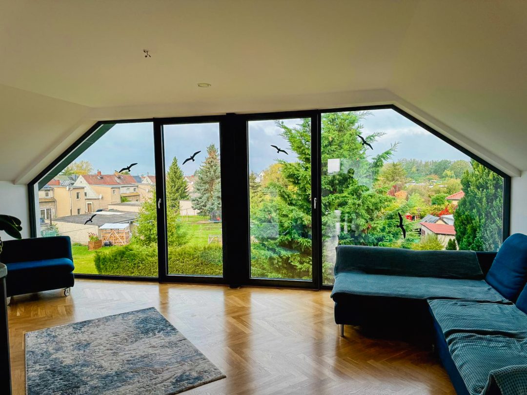 Die Innenaussicht eines Wohnzimmers durch eine große, bodentiefe, abgeschrägte Fensterfront, die den Blick auf einen grünen Garten und eine Siedlung freigibt. Im Raum sind ein blauer Ecksofa, ein kleinerer blauer Sessel und ein gemusterter Teppich auf einem Holzboden zu sehen. Schwarze Vogelsilhouetten sind dekorativ auf das Fensterglas geklebt.