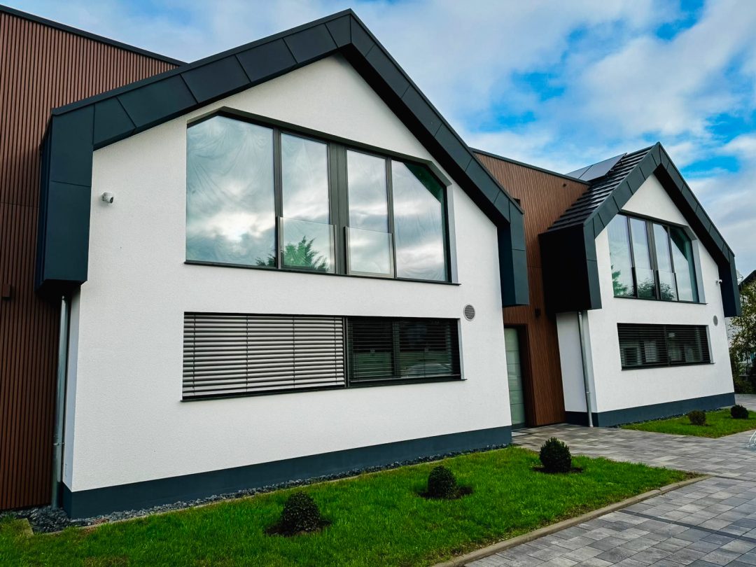 Nahaufnahme der weißen Putzfassade des modernen Doppelhauses. Die Fassade hat ein dunkles Giebeldach und große, dreieckige Fenster im Obergeschoss sowie ein rechteckiges Fenster mit Jalousien im Erdgeschoss. Links und rechts sieht man die braune Holzverkleidung der angrenzenden Gebäudeteile. Ein gepflasterter Weg führt auf das Haus zu, davor liegt ein grüner Rasen mit kleinen Büschen.