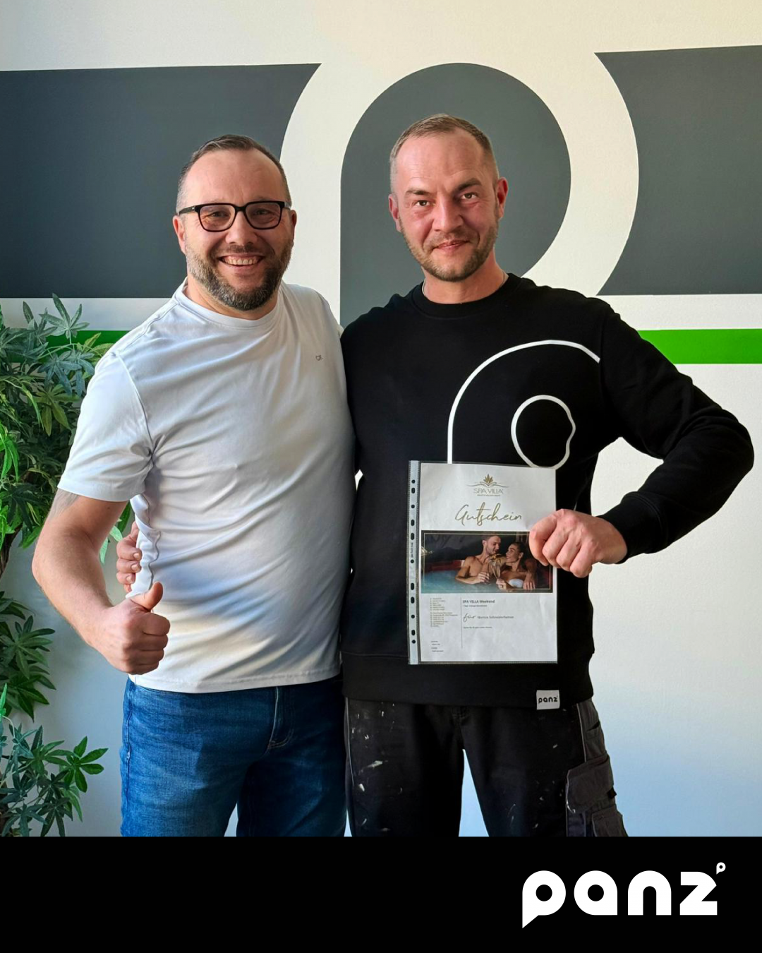 Lachende Männer im Büro der Panz Aluminiumtechnik GmbH. Der Mitarbeiter Markus (rechts, schwarzer Pullover) hält einen Gutschein für ein Wellnesswochenende in der Spa Villa Wingerode. Er wird vom Kollegen (links, weißes T-Shirt) für 20 Jahre Treue und Engagement gefeiert. Das Foto symbolisiert die Mitarbeiterwertschätzung und das Firmenjubiläum im Bereich Aluminiumtechnik und Metallbau.
