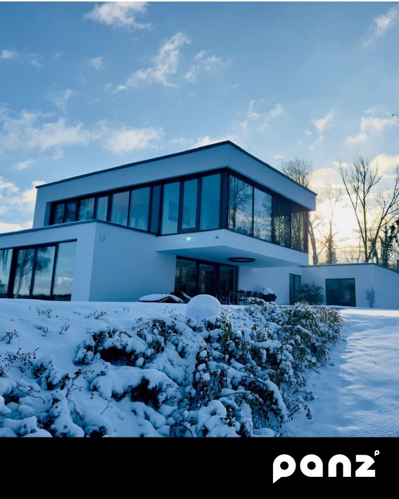 Modernes zweistöckiges Flachdachhaus in minimalistischer Architektur mit großen Glasfronten in einer verschneiten Winterlandschaft bei Sonnenschein.