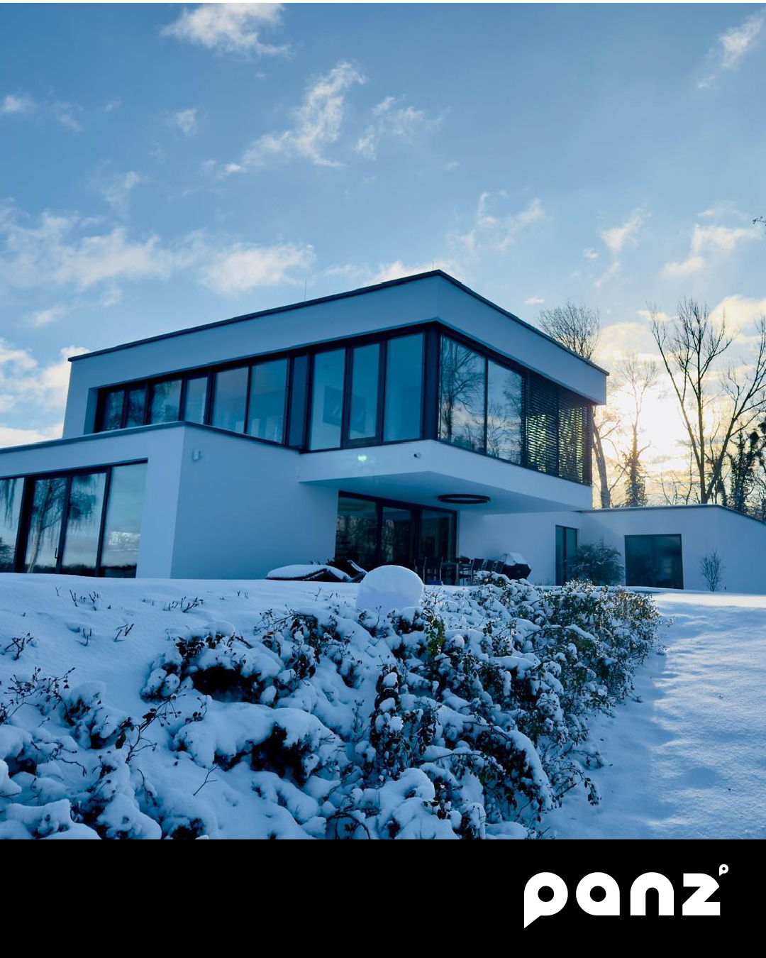 Modernes zweistöckiges Flachdachhaus in minimalistischer Architektur mit großen Glasfronten in einer verschneiten Winterlandschaft bei Sonnenschein.