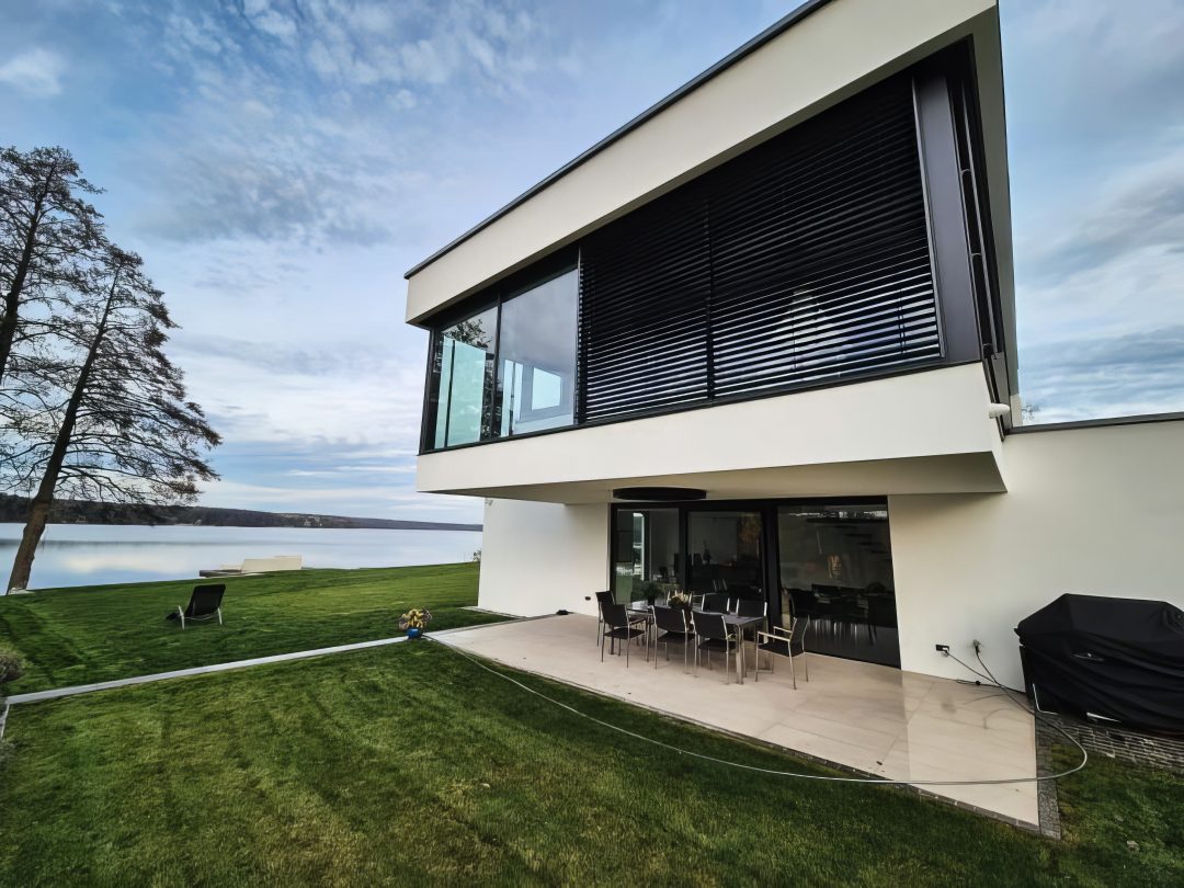 Moderne Architektur am See: Weiße Villa mit bodentiefen Schiebefenstern, Sonnenschutz-System und gepflegter Gartenanlage.