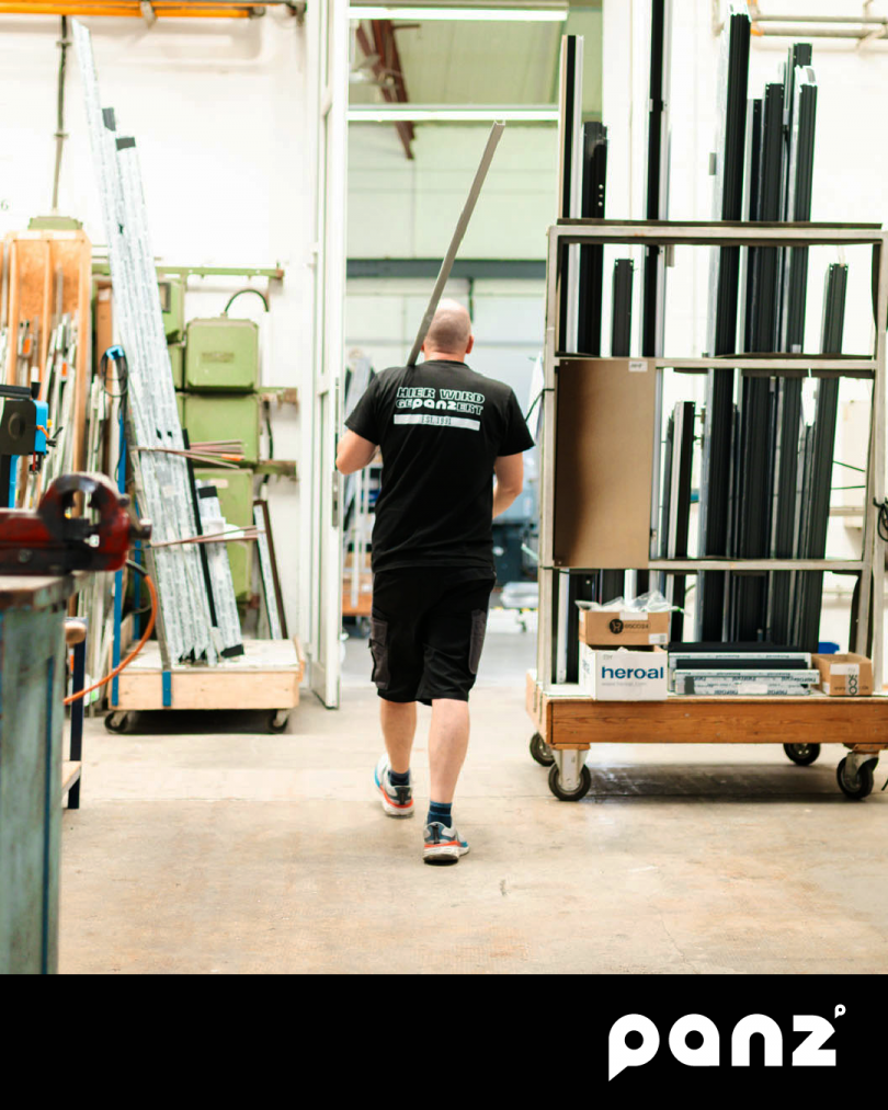 Mitarbeiter von Panz Aluminiumtechnik läuft mit einem Aluminiumprofil durch die Produktionshalle und wird von hinten fotografiert. Auf seinem T-Shirt steht "Hier wird gepanzert", unten rechts ist das panz-Logo zu sehen.