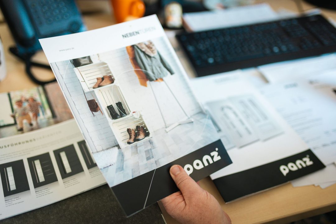 Eine Hand hält einen geöffneten Produktkatalog für Nebentüren der Marke Panz auf einem Schreibtisch im Büro. Im Hintergrund liegen weitere Broschüren und Büroutensilien wie eine Tastatur.