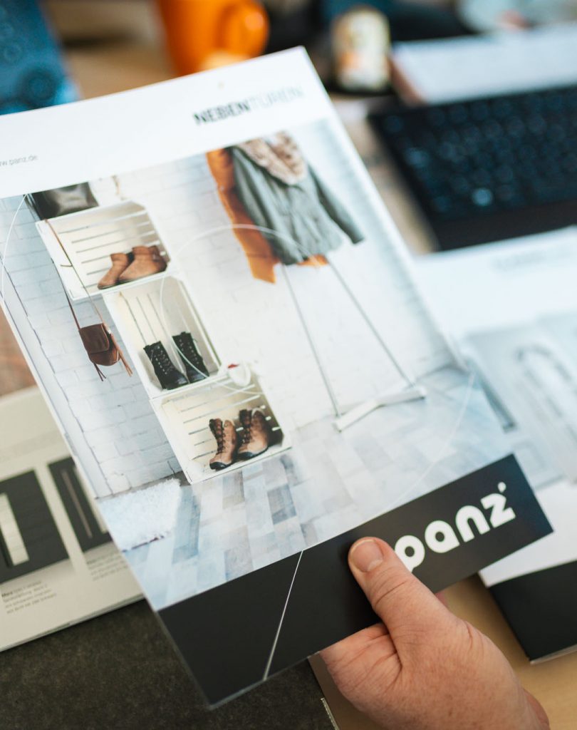 Eine Hand hält einen geöffneten Produktkatalog für Nebentüren der Marke Panz auf einem Schreibtisch im Büro. Im Hintergrund liegen weitere Broschüren und Büroutensilien wie eine Tastatur.