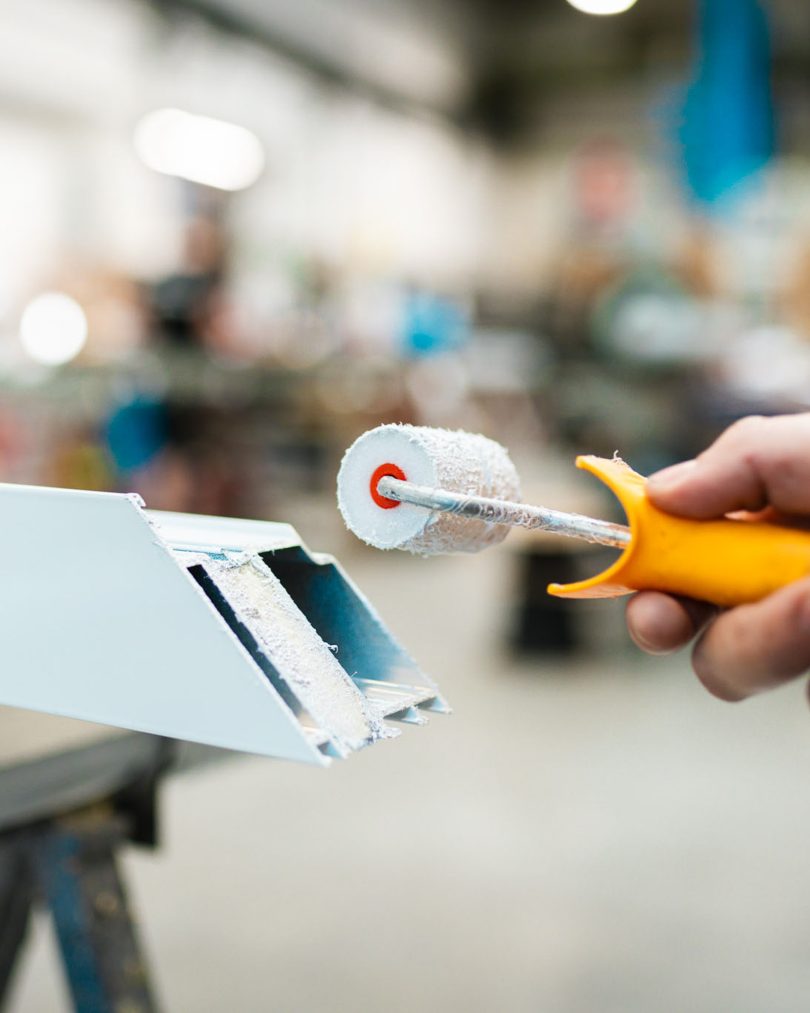 Nahaufnahme: Hand eines Handwerkers mit gelbem Griff führt kleine Lackierwalze an weißes Aluminiumprofil heran – Beschichtung und Oberflächenveredelung im Werk.
