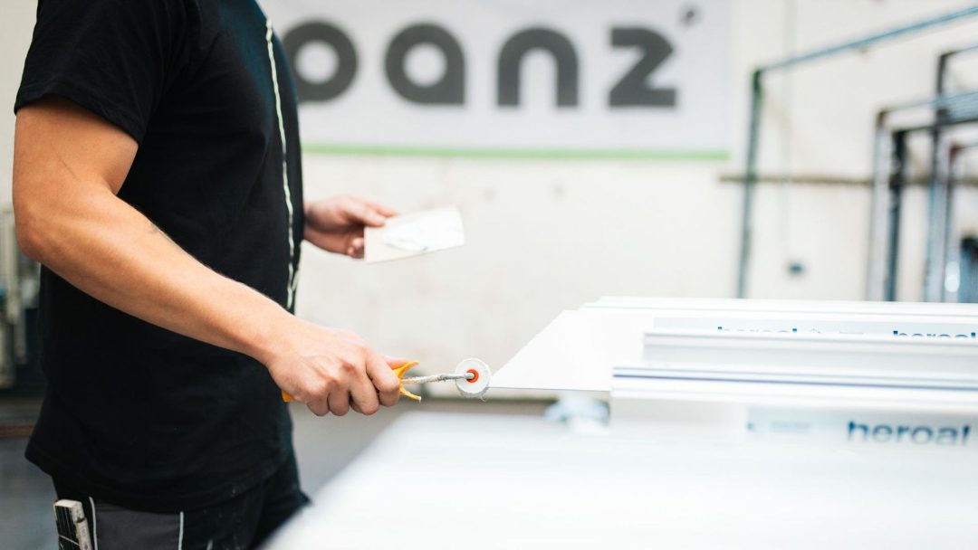 Handwerker in schwarzem Hemd mit Werkzeug und kleiner Schaumstoffrolle bei der Präzisionsarbeit an weißem Profil im Hintergrund 'panz' Banner.