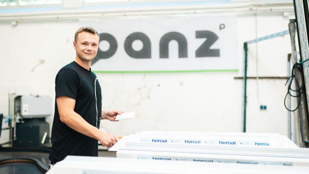 Gesamtansicht: Junger Handwerker lächelt in die Kamera, hält Werkzeug und kleines weißes Teil, blickt über mehrere gestapelte, beschichtete 'heroal' Aluminiumprofile – Werkstattumgebung.