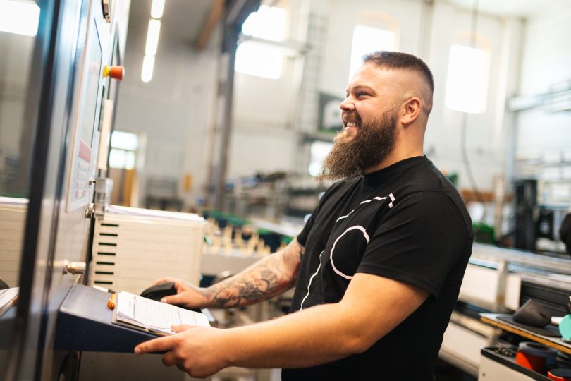 Ein tätowierter Arbeiter mit Bart bedient lächelnd das Steuerelement einer Produktionsanlage in einer Werkhalle